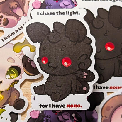 Black chibi Mothman sticker: He's sitting, with text that reads," I chase the light, for I have none".