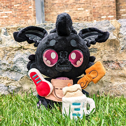 Sitting plush of Monty Mothman. A soft black and grey moth creature, with red eyes. Holding magnetic toy accessories for candle-making.